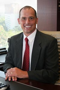 A photo of John Farley, McNICHOLS CFO and Senior VP, smiling at his desk.