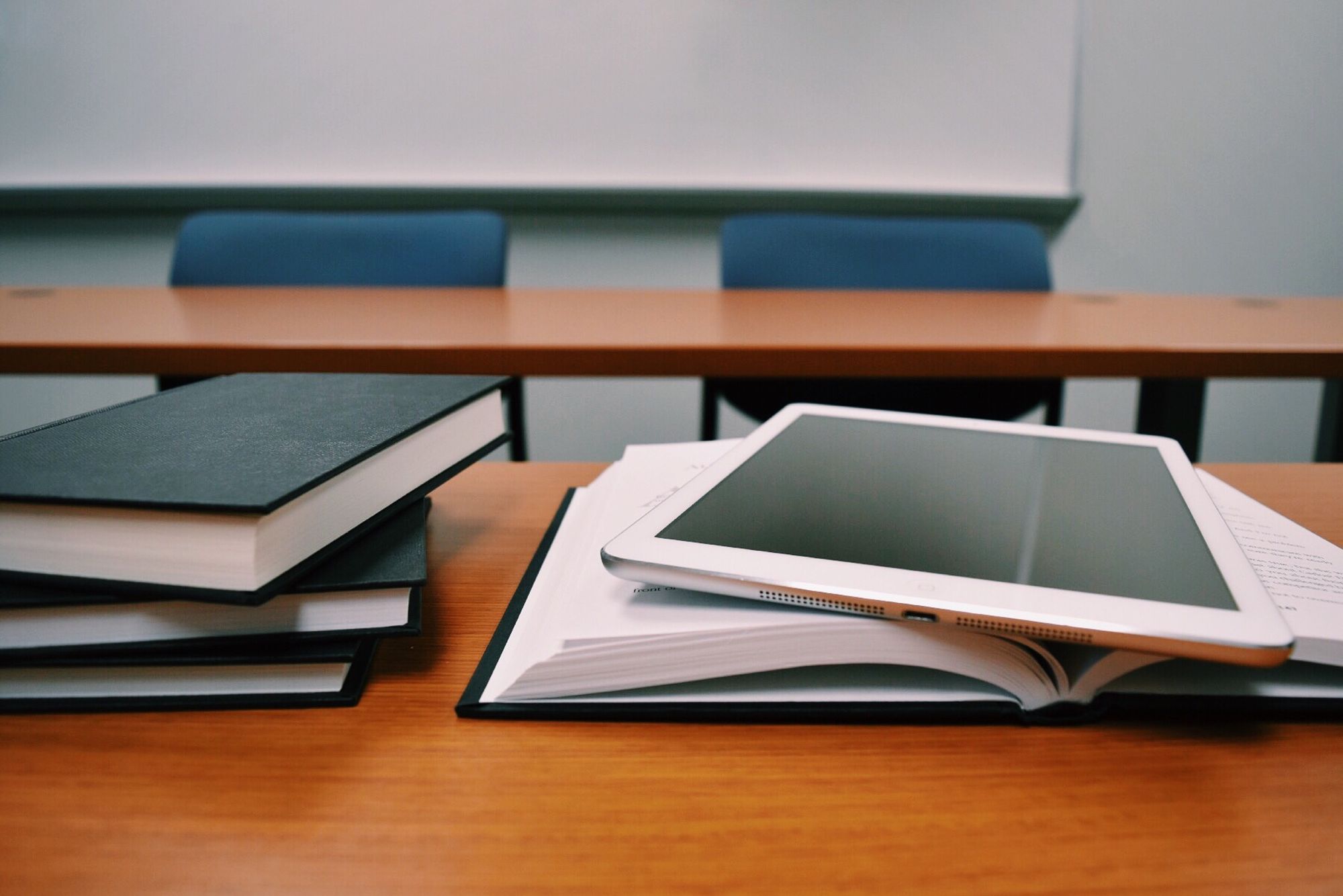 Stacks of books and an iPad on top of a wooden desk in a classroom.