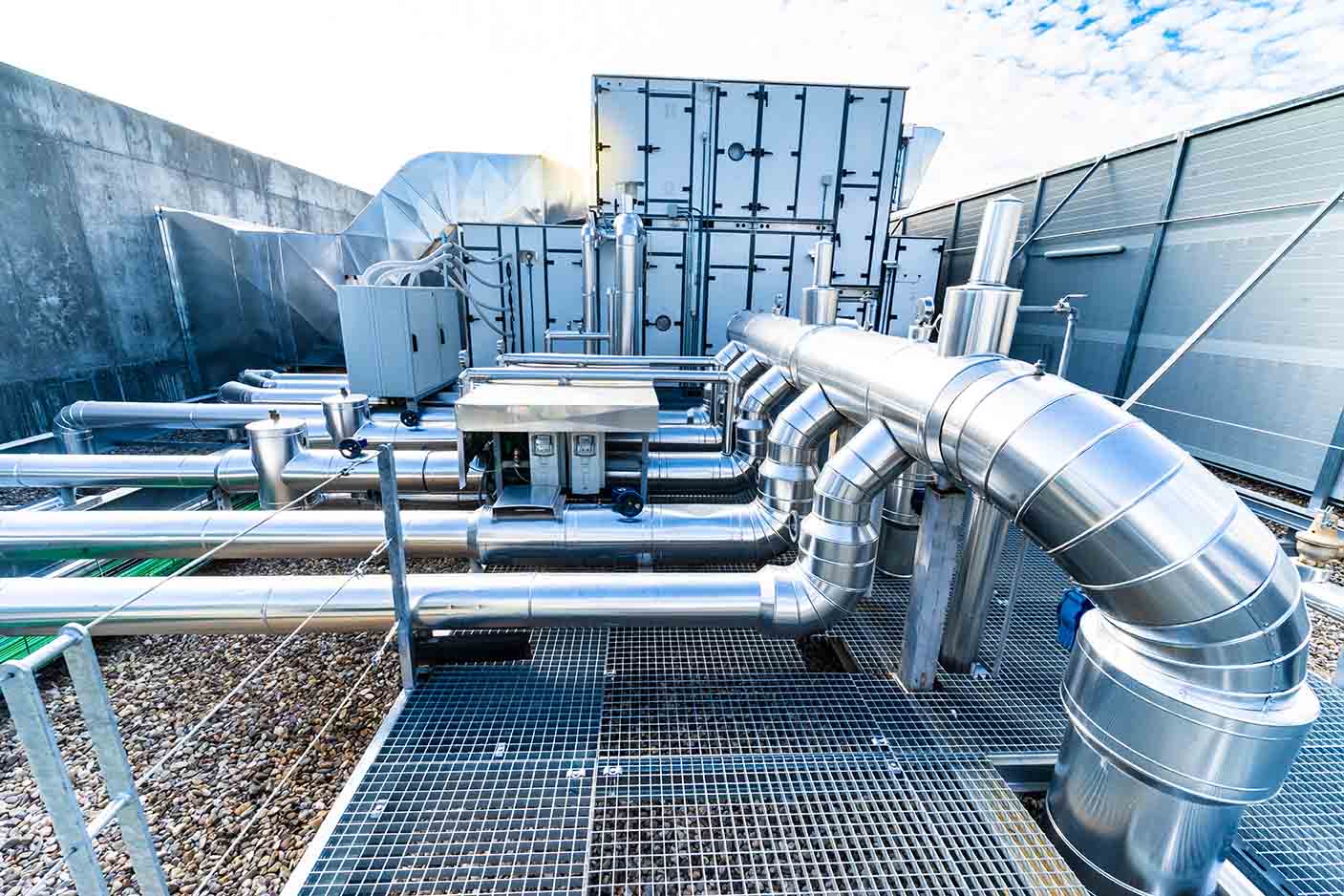 An industrial rooftop featuring stainless steel pipes and ducts connected to large HVAC units, supported by durable grated flooring, with a clear blue sky in the background.