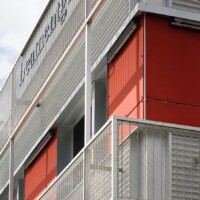 Building exterior with red panels and white framing, featuring Designer Mesh, AURA™ panels used as balcony and facade screening.