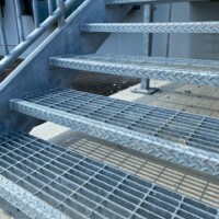 Smooth Standard-Duty Welded Bar Grating Stair Treads with serrated edges installed on a galvanized steel staircase with handrails.
