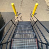 Smooth Standard-Duty Welded Bar Grating Stair Treads on an exterior metal staircase with handrails and yellow safety bollards at the base.